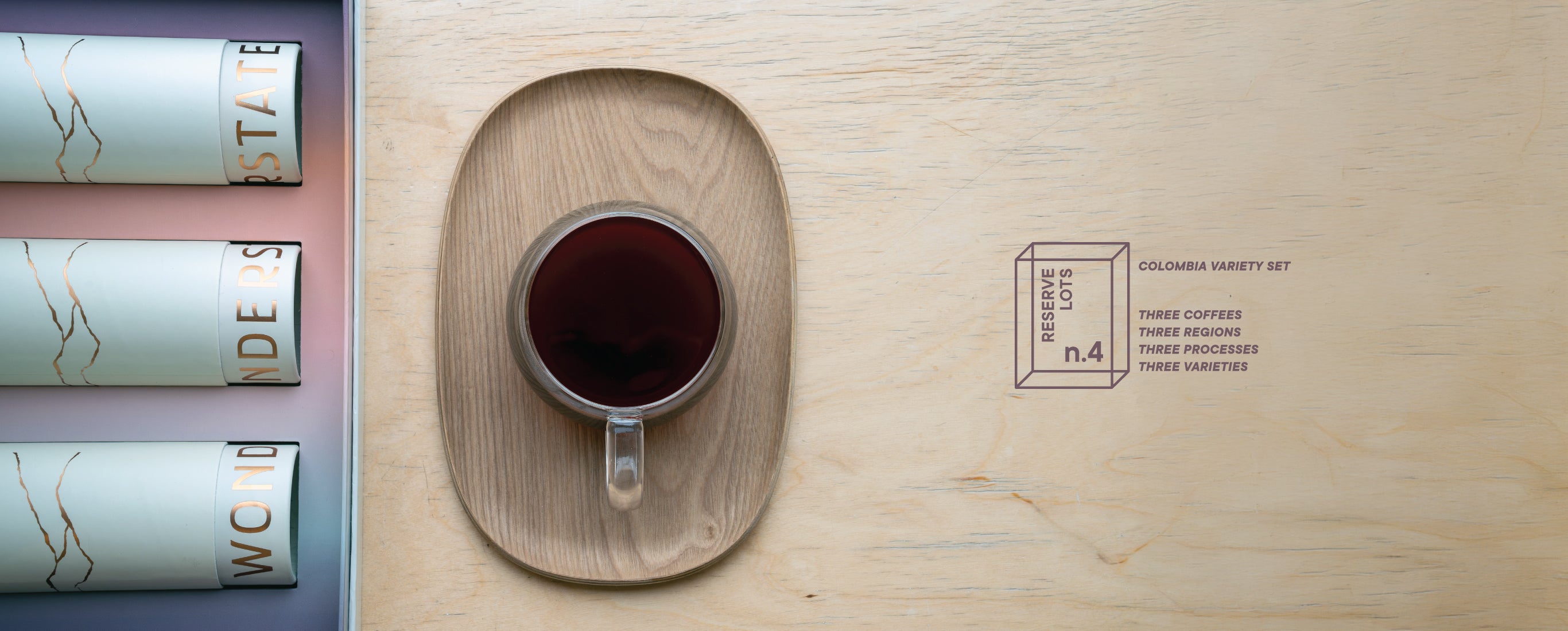 Cup of coffee on a wooden coaster with a branded box in the background. Colombia Variety Set Three Coffees Three Regions Three Processes Three Varieties