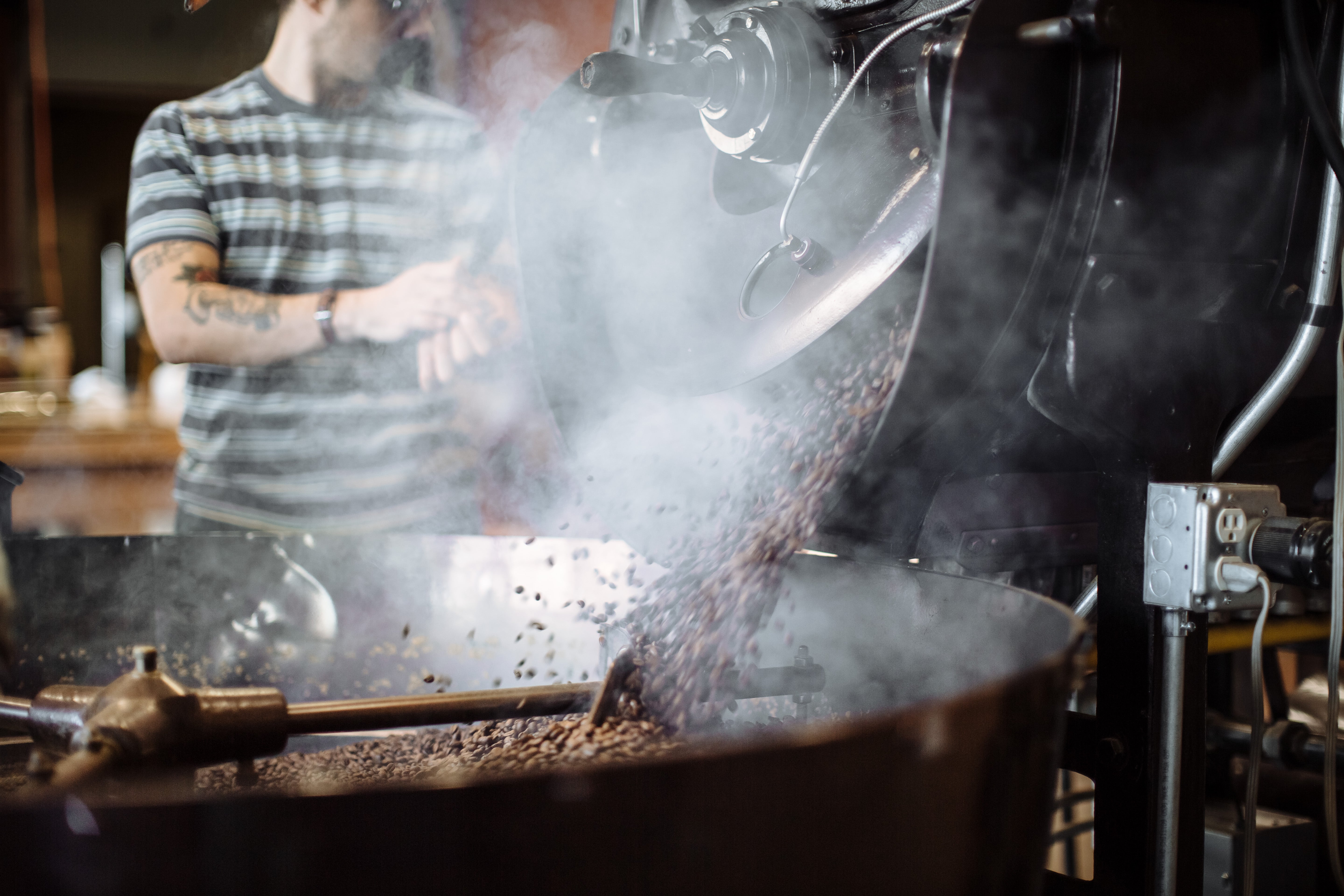Person operating a coffee roasting machine with smoke and coffee beans in the foreground