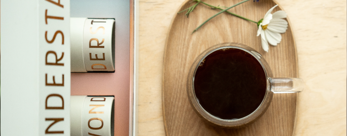 A top-down styled product shot showing a Wonderstate gift set box next to a glass cup of dark coffee on a wooden saucer, with a white flower and lavender sprig on a light wood tabletop.