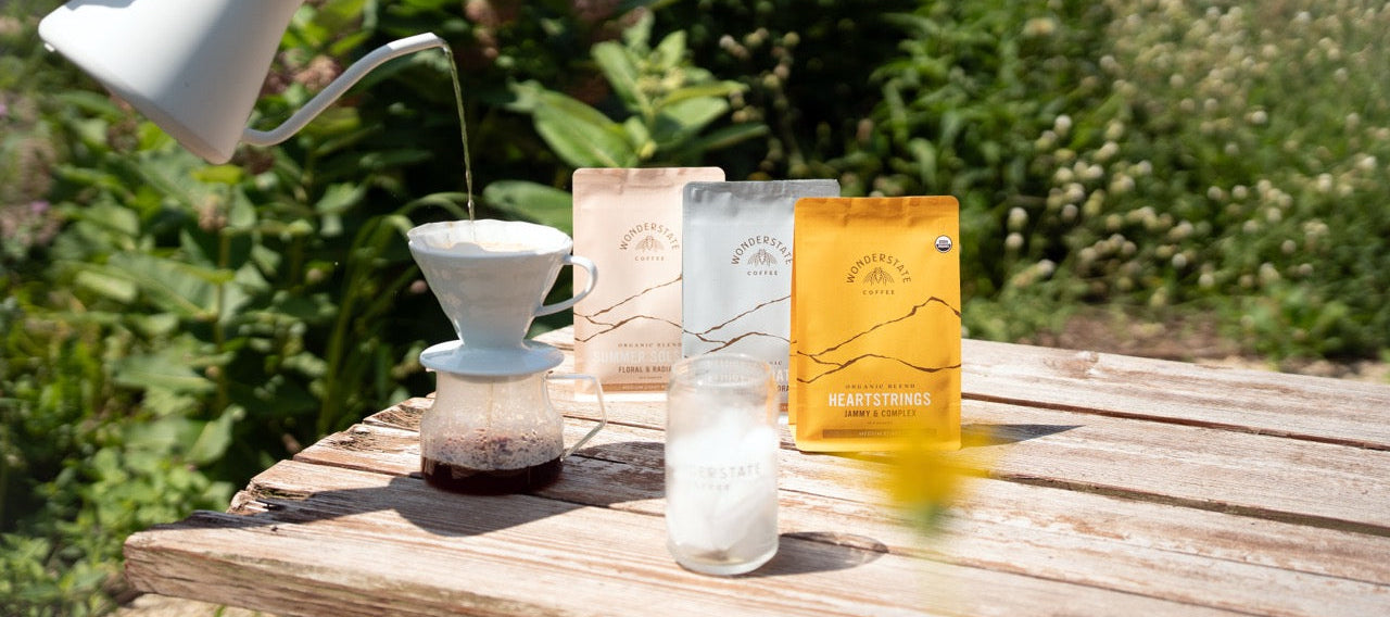 A hand pours hot water from a white pour-over coffee maker into a glass carafe on a wooden table outdoors. Three coffee bags from the brand Mountain Coffee Roasters are standing behind the setup, with a blurred garden background.