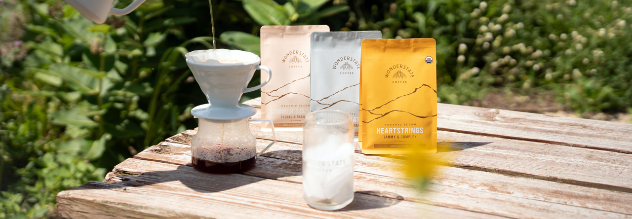 A hand pours hot water from a white pour-over coffee maker into a glass carafe on a wooden table outdoors. Three coffee bags from the brand Mountain Coffee Roasters are standing behind the setup, with a blurred garden background.