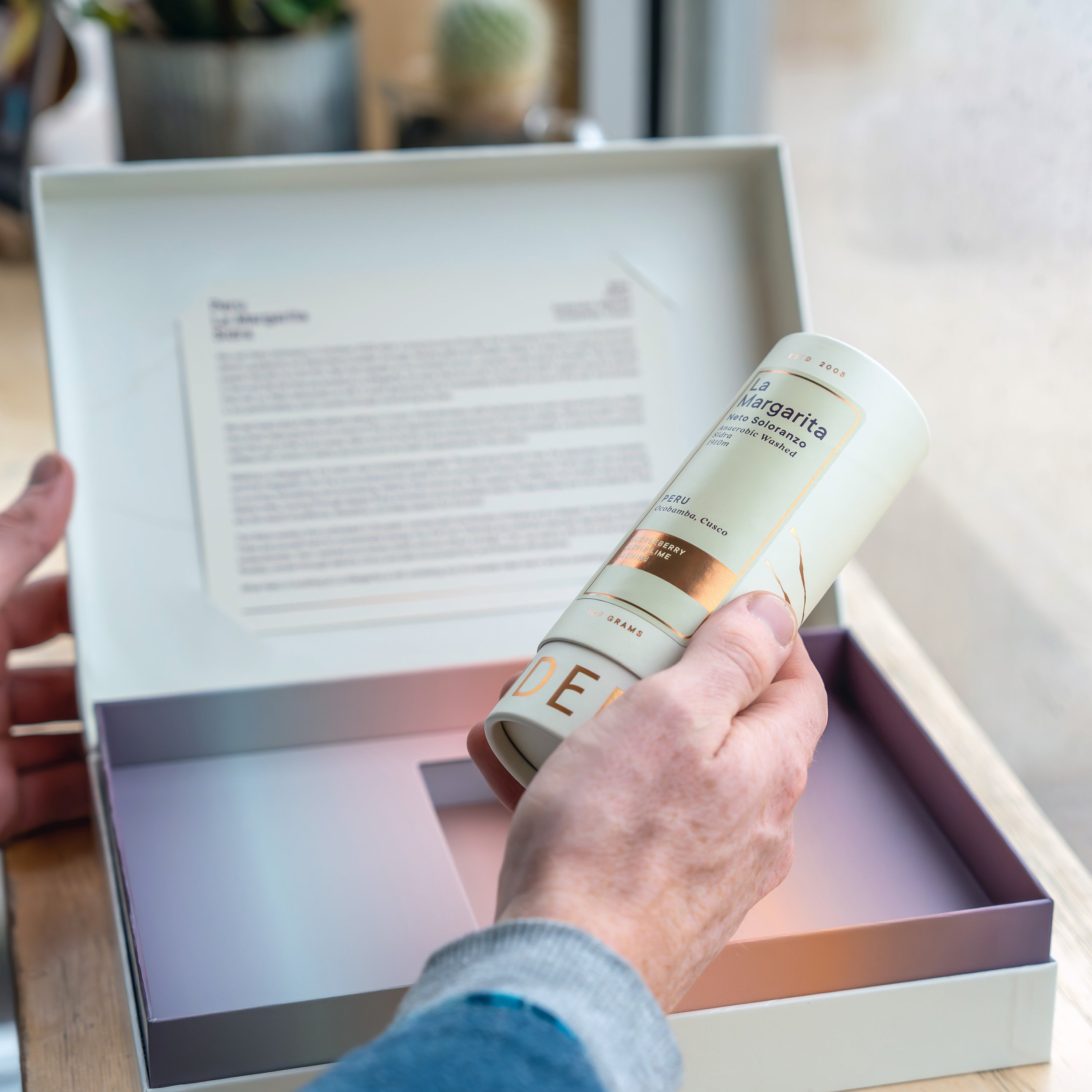 Person holding a cylindrical product labeled 'E' in front of an open box with printed material.